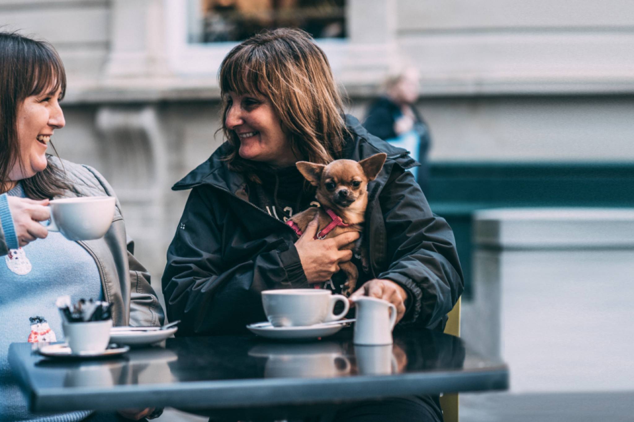 Two women are smiling and having coffee, and one is holding a small dog.
