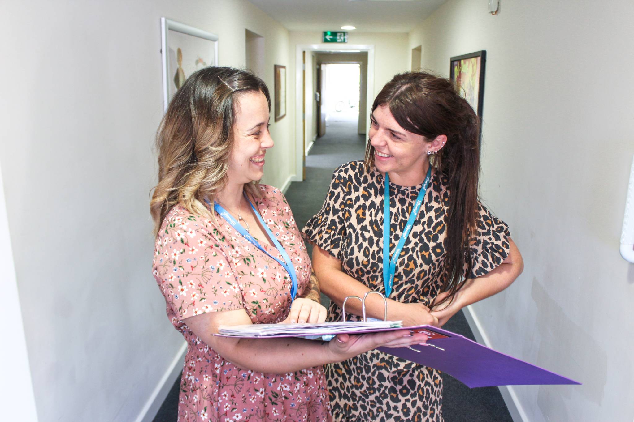 Two women stood in hallway looking over a document file, and smiling at each other